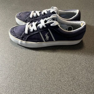 Mörkblå sneakers från Polo Ralph Lauren - Snygga mörkblå sneakers från Polo Ralph Lauren med vit logga på sidan och vita snören. Skorna har en klassisk låg profil, rund tå och vit sula med bruna detaljer undertill. Ovandelen är i canvas och insidan har mjuk textilkant.