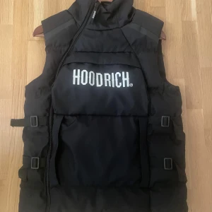 Svart dunväst från Hoodrich XS - Snygg svart dunväst från Hoodrich i storlek XS. Västen har stor vit logga framtill, justerbara spännen i sidorna och dragkedja med Hoodrich-detalj. Perfekt för lager-på-lager och streetwear-vibe.