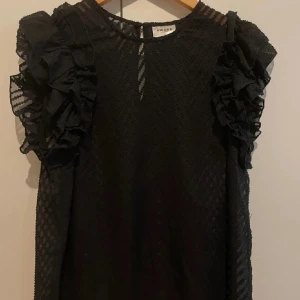 Svart volangklänning från Aware - Supersnygg svart kortklänning från Aware by Vero Moda med transparenta detaljer och volanger på axlarna. Klänningen har ett diskret mönster i tyget och är ärmlös med cool struktur. Perfekt för dig som gillar statement-ärmar och unik stil.
