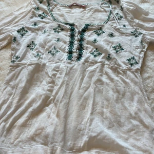 Vit blus med grön brodyr  - Supersöt vit blus med gröna broderade detaljer och dekorativa knappar framtill. Lätt och luftig bomullskänsla, långa ärmar med smock vid ärmslut. Perfekt för dig som gillar boho-vibe och unika detaljer.
