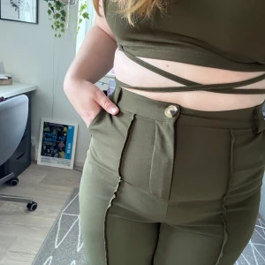 Olivgröna byxor med hög midja - Snygga olivgröna byxor med hög midja och vida ben. Byxorna har markerade sömmar framtill, fickor både fram och bak samt stängs med knapp och dragkedja. Sköna och stretchiga i materialet, perfekta för en trendig look.