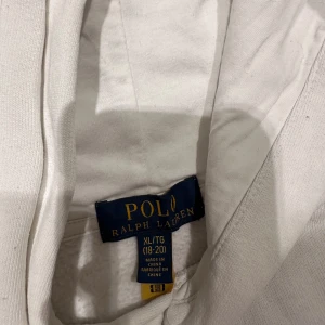 Vit hoodie Polo Ralph Lauren XL - Vit hoodie från Polo Ralph Lauren i storlek XL med broderad skidande björn på framsidan. Tröjan har huva och långa ärmar, tillverkad i mjuk bomull. Perfekt för dig som gillar klassisk stil med en twist och vill sticka ut med en ikonisk detalj.