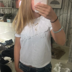 Vit blus med stor spetskrage - Säljer en vit blus med korta ärmar och en stor, dekorativ spetskrage med broderade detaljer. Blusen har en söt och unik look som sticker ut, perfekt att styla med jeans eller kjol. Materialet känns lätt och luftigt.
