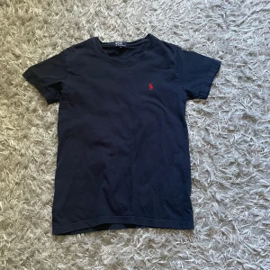 Svart t-shirt från Polo Ralph Lauren - Svart t-shirt från Polo Ralph Lauren i mjuk bomull. Klassisk passform med rund hals och korta ärmar. På bröstet finns den ikoniska röda Polo-loggan broderad. Perfekt för en clean och stilren look.
