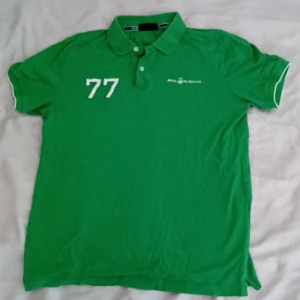 Grön pikétröja från Sail Racing - Snygg grön pikétröja från Sail Racing med vit broderad logga och siffran 77 på bröstet. Klassisk krage med knappar och korta ärmar. Perfekt för dig som gillar sportig och stilren look.