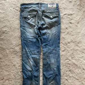 True Religion baggy jeans  - Säljer ett par blå baggy jeans från True Religion med ikoniska bakfickor och kontrastsömmar. Jeansen har klassisk femficksdesign, tydliga slitningar och detaljerade knappar. Perfekta för dig som gillar streetwear och vill sticka ut med snygga detaljer.