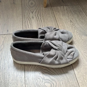Grå mocka slip-ons från Steve Madden - Gråa slip-on skor från Steve Madden i mjuk mocka med bred rosettdetalj framtill. Skorna har rund tå och vit platt sula, vilket ger en chill och trendig look. Perfekta att slänga på sig till jeans eller kjol för en avslappnad vibe.