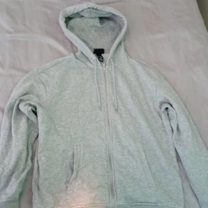 Ljusgrå hoodie med dragkedja - Enkel och stilren ljusgrå hoodie med dragkedja framtill och snörning i huvan. Tröjan har två fickor på sidorna och är tillverkad i mjuk bomullsblandning. Perfekt för chill dagar eller när du vill ha en clean look.