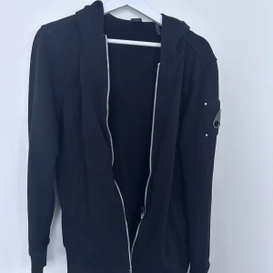 Svart hoodie från Moose Knuckles 💫 - Svart hoodie från Moose Knuckles med dragkedja hela vägen, huva och snygg metallogo på ärmen. Tröjan har långa ärmar, två fickor fram och en clean, stilren look som passar perfekt till streetwear. Materialet är mjuk bomull.