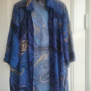 Blå paisleymönstrad blus MC Fashion - Säljer en luftig lång blus från MC Fashion med paisleymönster i olika blå nyanser och inslag av beige. Blus har trekvartsärm, skjortkrage och är tillverkad i ett tunt, syntetiskt material. Perfekt för dig som gillar färg och mönster!
