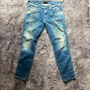 Blå Replay Anbass jeans herr - Säljer ett par blå Replay Anbass jeans med snyggt slitna detaljer och klassisk femficksdesign. Jeansen har smal passform och raka ben, tillverkade i mjukt bomullsmaterial med lite stretch för extra komfort. Perfekta för dig som gillar en modern och avslappnad stil.