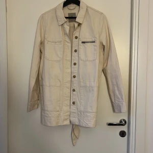 Beige overshirt från Vero Moda - Snygg beige overshirt från Vero Moda med knappar framtill, flera fickor och ett bälte i midjan. Jackan har klassisk krage och långa ärmar, perfekt för lager-på-lager. Enkel och clean stil som passar till det mesta.
