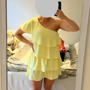 Gul omlottklänning med volanger från Zara - Säljer en ljusgul kort klänning från Zara i storlek M. Klänningen har ett snyggt oneshoulder-snitt och flera lager med volanger. Det finns en svag obehandlad fläck under ena volangen. Der är ingen som syns när man har klänningen på sig. 