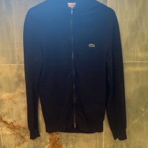 Mörkblå kofta från Lacoste fullzip  - Snygg mörkblå kofta från Lacoste med dragkedja framtill och klassisk krokodil-logga på bröstet. Regular fit och långärmad modell, perfekt för lager-på-lager. Tillverkad i mjukt bomullsmaterial som känns skönt mot huden.