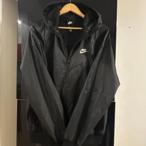 Svart vindjacka från Nike med huva - Svart oanvänd vindjacka från Nike med klassisk vit logga på bröstet. Jackan är i storlek M men lite stor i storlek, därav har den ej kommit till användning. 