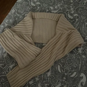 Beige ribbad stickad bolero - Supersnygg beige stickad bolero med ribbad struktur. Perfekt att slänga över en topp för en trendig lager-på-lager-look. Mjuk och skön, med långa ärmar och bred krage. Passar dig som gillar stilrena och neutrala färger.