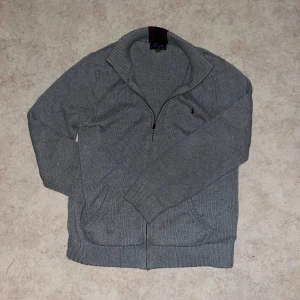 Grå stickad kofta från Polo Ralph Lauren - Snygg grå stickad kofta från Polo Ralph Lauren med dragkedja framtill och två fickor. Klassisk design med ribbade muddar och diskret logga på bröstet. Perfekt för lager-på-lager och chill vardagsstil.