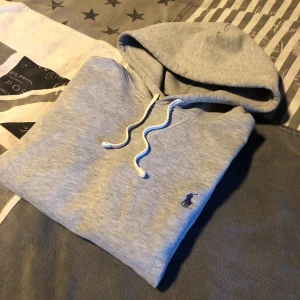 Grå hoodie från Polo Ralph Lauren - Snygg grå hoodie från Polo Ralph Lauren med klassisk blå logga på bröstet. Tröjan har huva med snörning, stor magficka och långa ärmar. Perfekt för chill dagar och enkel att matcha med jeans eller joggers. Mjuk bomullskvalitet som känns skön mot huden.