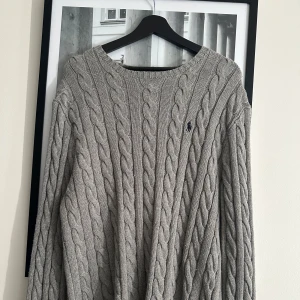 Grå kabelstickad tröja Polo Ralph Lauren - Kabelstickad grå tröja från Polo Ralph Lauren med klassisk rund hals och broderad logga på bröstet. Tillverkad i 100% bomull och har långa ärmar samt ribbade muddar. Perfekt för lager-på-lager och snygg till jeans eller kjol.