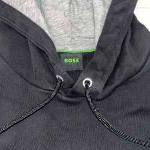 Svart hoodie från BOSS med broderad logga - Svart hoodie från BOSS med stor broderad logga på bröstet. Tröjan har huva med snörning, känguruficka och mjukt grått innerfoder i luvan. Perfekt för en chill och stilren look.