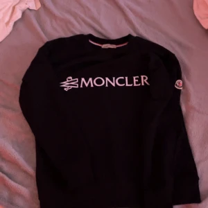 Svart Moncler sweatshirt med logga S passar kan passa M - Svart sweatshirt från Moncler med stor vit logga framtill och klassisk Moncler-patch på ärmen. Tröjan har rund halsringning, ribbade muddar och är långärmad. Perfekt för dig som gillar stilrena och exklusiva plagg.