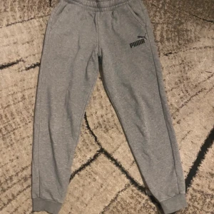 Grå mjukisbyxor från Puma - Snygga grå mjukisbyxor från Puma med svart logga på vänster sida. Byxorna har elastisk midja, ribbade muddar vid benslut och två sidofickor. Perfekta för chill eller träning, tillverkade i mjukt bomullsmaterial för extra komfort.
