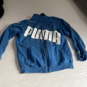 Blå ziptröja från Puma med logga - Blå ziptröja från Puma med stor vit logga över bröstet. Tröjan har hel dragkedja, ståkrage och två fickor framtill. Tillverkad i mjuk bomullsblandning, perfekt för chill eller träning. Klassisk sportig look med ribbade muddar och detaljer vid ärmslut.