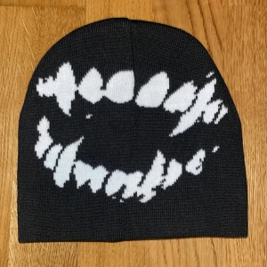 Svart mössa med vit tandmönster - Cool svart mössa med ett stort vitt tandavtrycksmönster framtill. Mössan är stickad och har en klassisk passform som sitter snyggt på huvudet. Perfekt för dig som vill sticka ut med en edgy och unik accessoar.