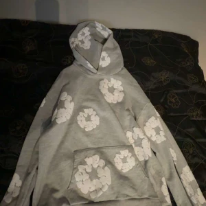 Grå hoodie från denim tears - Säljer en grå hoodie med stora vita blommönster över hela tröjan. Tröjan har huva och en stor magficka framtill. Perfekt för dig som gillar en chill och trendig stil. Passar både till jeans och mjukisbyxor. Står lx men passar både L och XL!