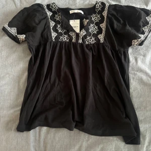 Svart blus med broderade detaljer - Svart blus från Spanien med vita broderade mönster längs halsringningen och på ärmarna. Blusen har korta puffärmar och en lös passform, perfekt för en chill och stilren look. Materialet känns mjukt och luftigt, och de broderade detaljerna ger en unik touch.