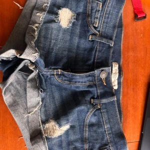 Jeansshorts från Abercrombie & Fitch - Snygga blå jeansshorts från Abercrombie & Fitch med slitna detaljer och råa kanter. Klassisk femficksmodell med coola slitningar framtill och dekorativa sömmar på bakfickorna. Perfekta för sommardagar och festivalhäng.