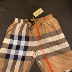Burberry shorts - Snygga shorts från Burberry med klassiskt rutigt mönster i beige, svart, vit, grå och röd. De har elastisk midja med svart snörning och är tillverkade i ett lätt, syntetiskt material.