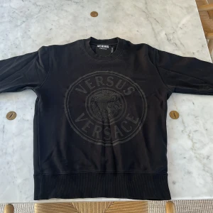 Svart sweatshirt Versus Versace S - Svart sweatshirt från Versus Versace i storlek S. Tröjan har ett stort, ton-i-ton tryck med Versus Versace-loggan och ett lejonmotiv på bröstet. Rund halsringning och ribbade muddar vid ärmslut och nederkant. Perfekt för dig som gillar stilrena designer med lyxig känsla.