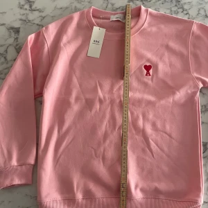 Rosa sweatshirt från Ami - Snygg rosa sweatshirt från Ami Alexandre Mattiussi med klassisk rund hals och ribbade muddar. På bröstet finns ett rött broderat hjärta med bokstaven A. Tröjan är i mjuk bomull och har lång ärm, perfekt för dig som gillar stilrena och coola detaljer. Stolek xs, helt oanvänd