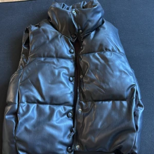 Svart dunväst i skinnimitation från Zara - Säljer en svart, quiltad dunväst från Zara i skinnimitation. Västen har hög krage, dragkedja och tryckknappar framtill och fickor på båda sidorna. Perfekt för lager-på-lager och ger en cool vibe till din outfit.