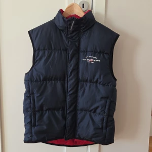 Mörkblå dunväst Polo Jeans Ralph Lauren - Snygg mörkblå dunväst från Polo Jeans Ralph Lauren med broderad logga på bröstet. Västen har hög krage, två fickor med tryckknapp och röd insida. Yttermaterial i polyester och fyllning av dun och fjäder. Perfekt lager-på-lager-plagg för kyliga dagar.