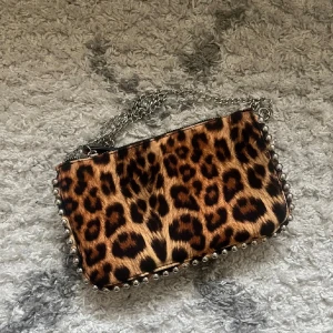 Leopardmönstrad axelväska med kedja - Cool axelväska med leopardmönster och silverfärgad kedja. Väskan har dekorativa silverkulor längs kanterna och dragkedja upptill. Perfekt för dig som vill sticka ut med en trendig accessoar. Köpt från zara för 300kr, helt oanvänd men har tyvärr inte prislappen kvar