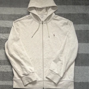 Beige hoodie från Polo Ralph Lauren - Snygg beige hoodie från Polo Ralph Lauren med dragkedja och huva. Klassisk broderad logga på bröstet och praktiska fickor framtill. Tillverkad i mjuk bomullsmix, perfekt för chill dagar. Långärmad och med snörning i huvan för extra stilpoäng. 