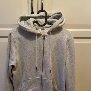 Ljusgrå hoodie med dragkedja H&M - Ljusgrå hoodie från H&M i storlek S. Tröjan har huva med snörning, hel dragkedja framtill och två fickor. Perfekt för chill dagar eller när du vill ha en enkel och clean look. Tillverkad i mjuk bomullsblandning.