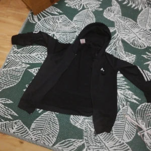 Svart hoodie från Adidas med huva - Svart hoodie från Adidas med dragkedja och huva. Klassiska tre ränder på ärmarna och liten Adidas-logga på bröstet. Långärmad modell i mjukt material, perfekt för chill eller träning.