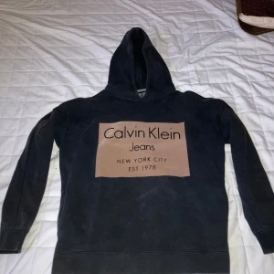 Svart hoodie Calvin Klein Jeans S - Svart hoodie från Calvin Klein Jeans med stor logga i beige framtill. Klassisk modell med huva och långa ärmar. Mjuk insida och ribbade muddar. Perfekt för en chill och stilren look.