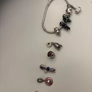 Silverfärgat armband med Marvel-berlocker - Säljer ett coolt silverfärgat armband med flera Marvel- och Star Wars-inspirerade berlocker, bland annat Captain Americas sköld, Thors hammare, Iron Man, Hulk, Black Panther, Spiderman och R2-D2. Några berlocker har färgdetaljer i rött, blått, grönt och guld. Perfekt för dig som älskar superhjältar och nördiga accessoarer. 1 st  pris 200 kr allt med 1800 kr