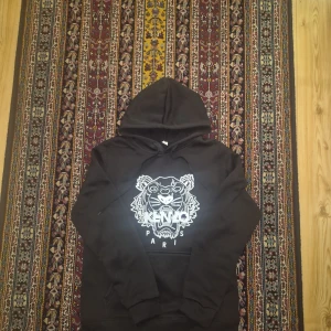 Svart Kenzo hoodie med tigertryck - Svart hoodie från Kenzo med ikoniskt vitt tigertryck och texten 'KENZO PARIS' på bröstet. Klassisk huva med snörning och känguruficka framtill. Perfekt för dig som gillar streetstyle och vill sticka ut med en fet logga.