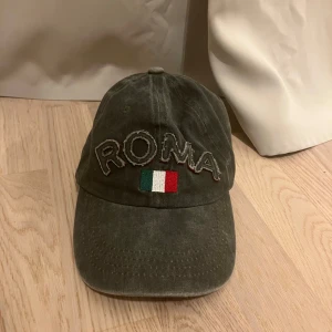 Olivgrön keps ROMA Italia - Olivgrön keps med broderad texten ROMA och Italiens flagga framtill. Från en marknad i Rom.  Justerbar baktill för perfekt passform. Tillverkad i 100% bomull, perfekt för dig som gillar italiensk stil och streetwear-vibe.