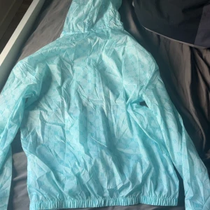 Turkos vindjacka från JGHFL - Säljer en fräsch turkos vindjacka från JGHFL med mönstrade B-loggor över hela jackan. Jackan har huva med orange snörning, dragkedja framtill och ett orange patch på ärmen. Perfekt för dig som gillar streetwear och vill sticka ut med färg.