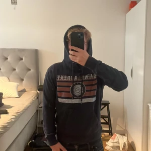 Saint Laurent marinblå hoodie med tryck - Säljer en sällsynt marinblå hoodie från Saint Laurent med stort tryck på bröstet i rött, vitt och blått. Tröjan har huva med snörning och en klassisk känguruficka framtill. Perfekt för dig som gillar streetwear och snygga detaljer. Storlek S passar 165-170, kan även ta kort på lappen på insidan, den är skön och fräsch, har växt ur hoodien nu efter dom här två åren, ställ frågor så svarar jag😊 Priset är inte hugget i sten så ge prisförslag den som budar högst eller köper direkt för den🥰