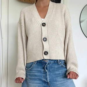 Beige ribbad kofta från H&M - Säljer en beige ribbad kofta från H&M med stora svarta och bruna knappar framtill. Koftan har v-ringad hals, långärmade ärmar. Perfekt för höst och vintern