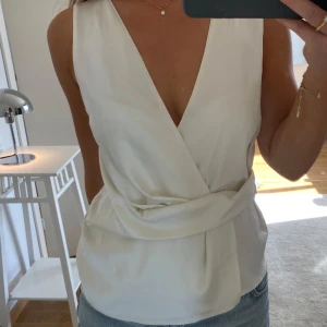 Vit omlottopp med djup v-ringning - Säljer en stilren vit omlottopp med djup v-ringning och bred knytdetalj i midjan. Toppen är ärmlös och har en elegant, enkel look som passar perfekt till jeans eller kjol. Materialet känns mjukt och har ett lätt glansigt utseende.