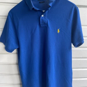 Blå Ralph Lauren pikétröja - Klassisk blå pikétröja från Ralph Lauren med gul broderad logga på bröstet. Tröjan har korta ärmar, krage och knäppning med två vita knappar. Tillverkad i 100% bomull för en skön och luftig känsla. Perfekt för en clean och sportig stil.