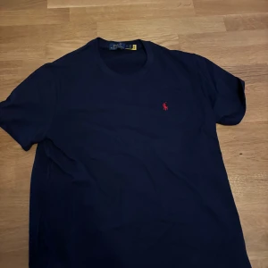 Mörkblå t-shirt från Polo Ralph Lauren - Snygg mörkblå t-shirt från Polo Ralph Lauren med klassisk röd logobrodyr på bröstet. Modellen är kortärmad och har en rund hals. Tillverkad i mjuk bomull som känns skön mot huden. Perfekt för dig som gillar stilrena och tidlösa plagg.